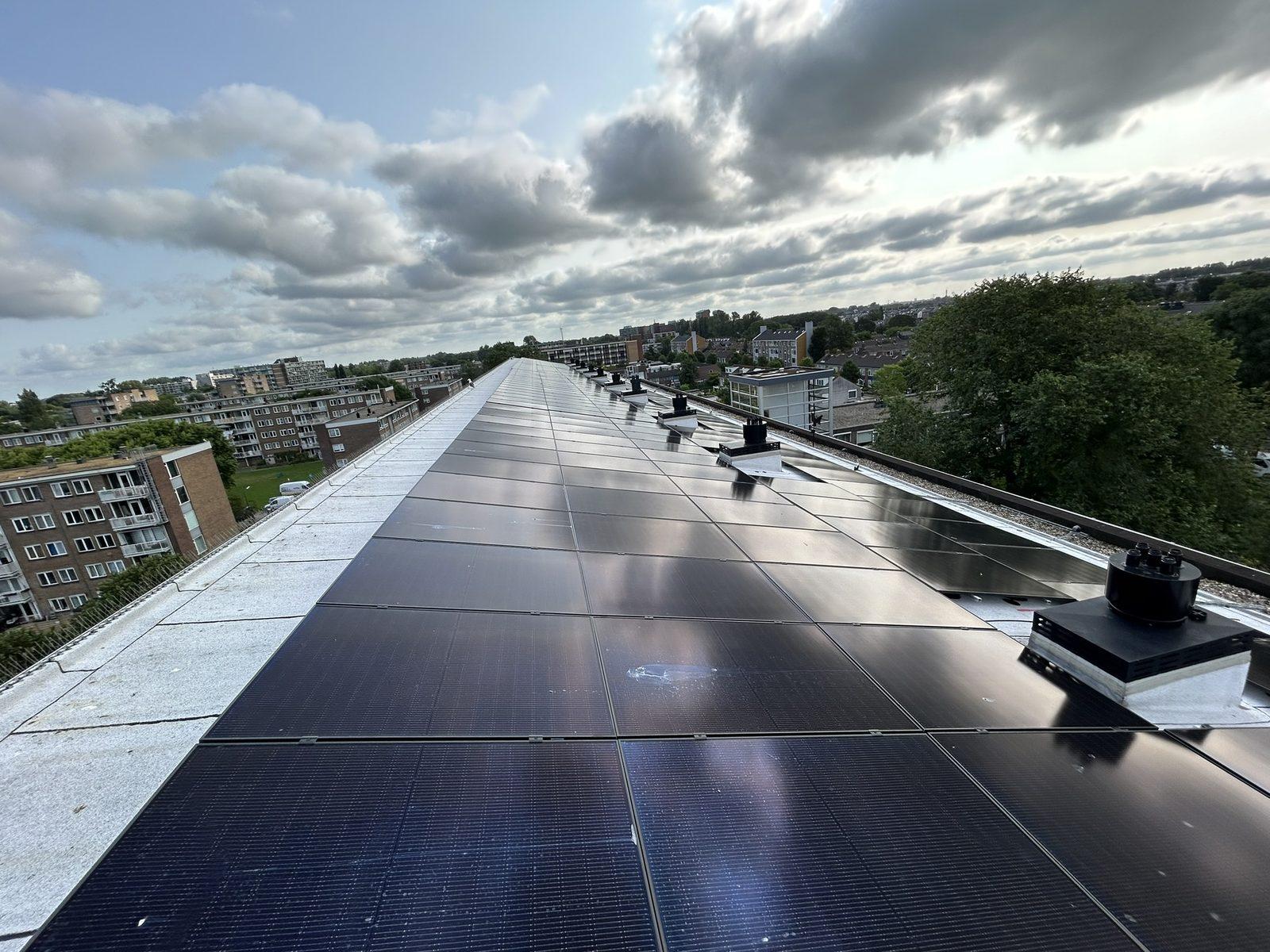 Lange rij zonnepanelen over het volledige platte dak van een VvE-flat, met uitzicht over de omliggende woonwijk
